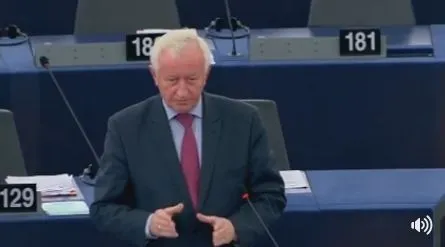 Starszy mężczyzna w garniturze i czerwonym krawacie przemawia przy mównicy w sali obrad z numerowanymi miejscami.