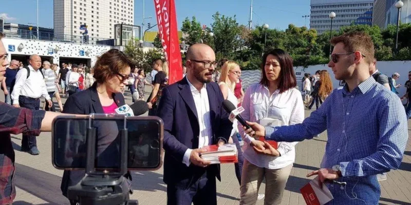 Mężczyzna w garniturze trzyma stos dokumentów o konstytucji i udziela wywiadu dziennikarzom z mikrofonami i kamerą na miejskim placu.