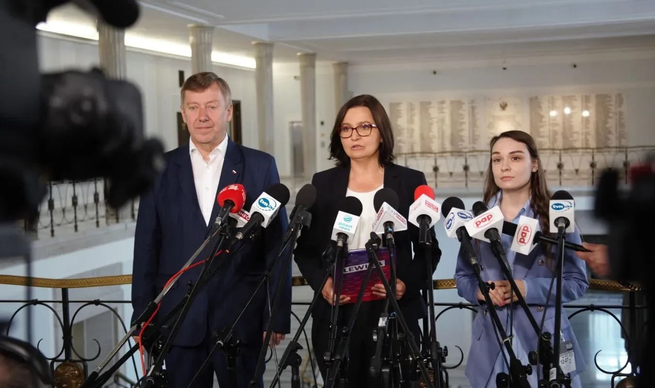 Trzy osoby stoją w eleganckim holu przy licznych mikrofonach podczas konferencji prasowej, omawiając temat niedoborów witaminy D.