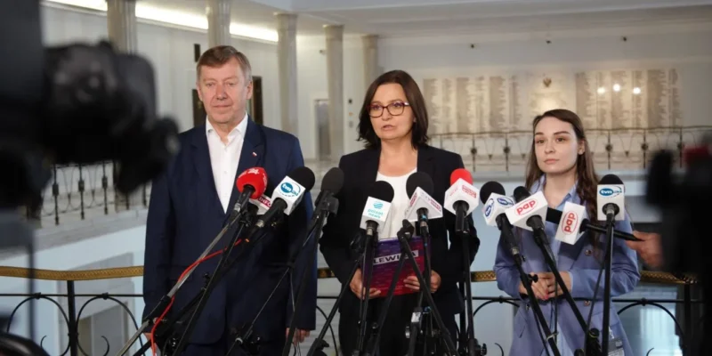 Trzy osoby stoją w eleganckim holu przy licznych mikrofonach podczas konferencji prasowej, omawiając temat niedoborów witaminy D.