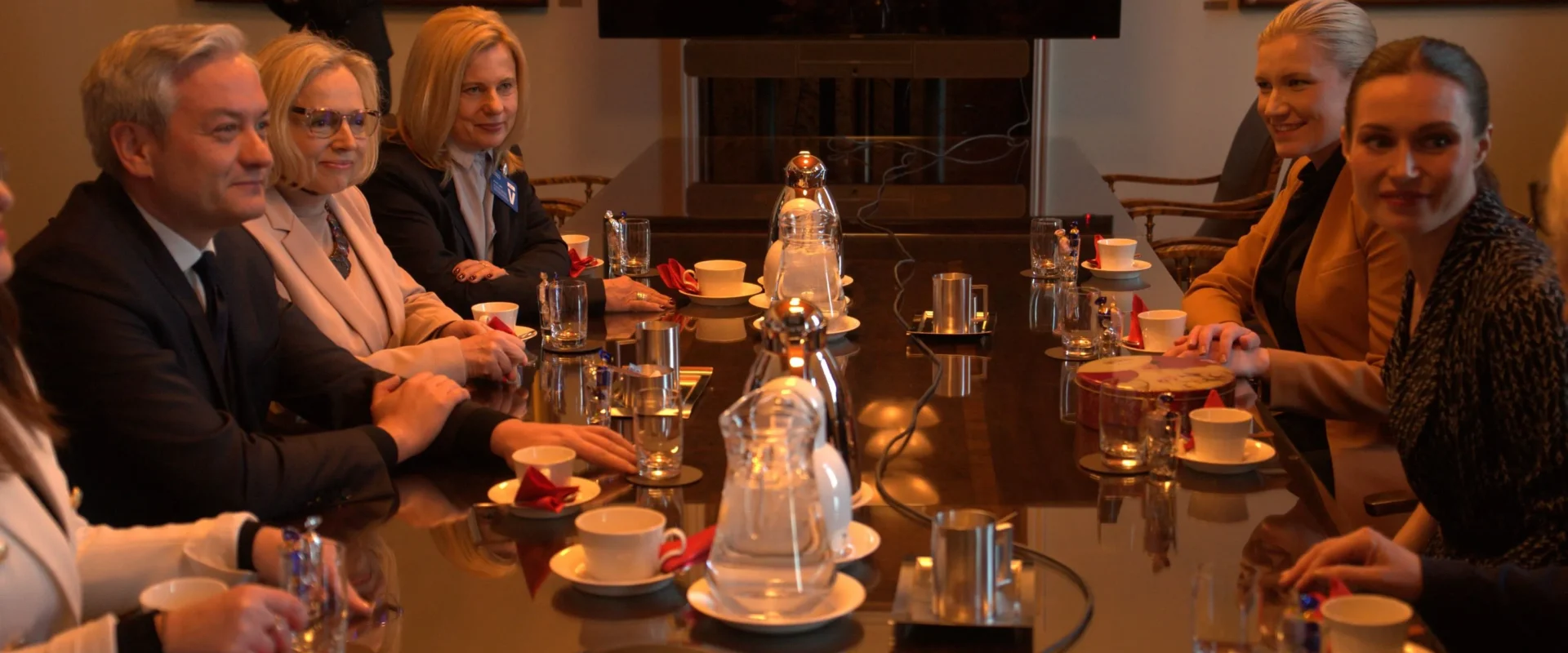 Na zdjęciu widać grupę osób siedzących przy stole konferencyjnym, rozmawiających w eleganckim otoczeniu.