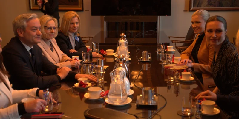 Na zdjęciu widać grupę osób siedzących przy stole konferencyjnym, rozmawiających w eleganckim otoczeniu.