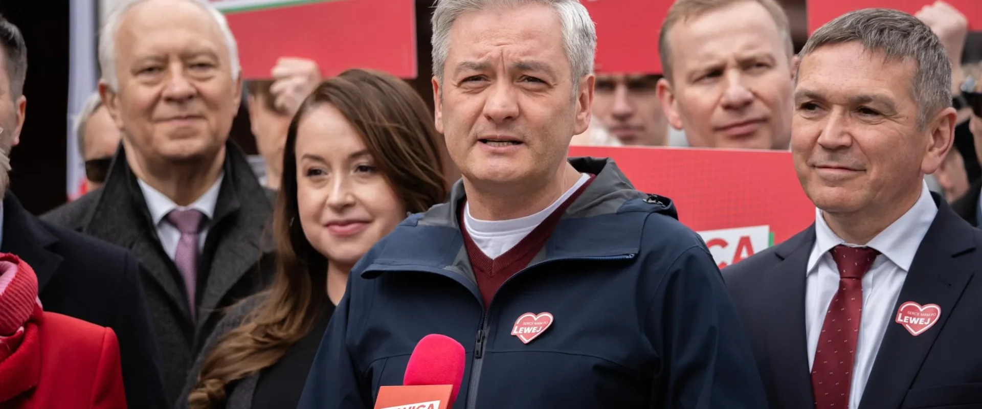 Grupa osób podczas wydarzenia Lewicy, na pierwszym planie mężczyzna w granatowej kurtce przemawia do mikrofonu, w tle czerwone plakaty z napisem LEWICA.