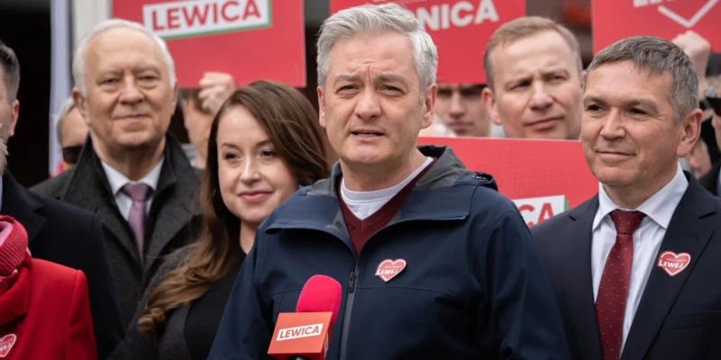 Grupa osób podczas wydarzenia Lewicy, na pierwszym planie mężczyzna w granatowej kurtce przemawia do mikrofonu, w tle czerwone plakaty z napisem LEWICA.