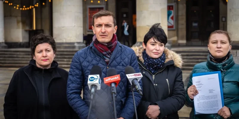 Cztery osoby stoją przed wejściem do budynku i udzielają konferencji prasowej z kilkoma mikrofonami, a jedna z nich trzyma dokument.