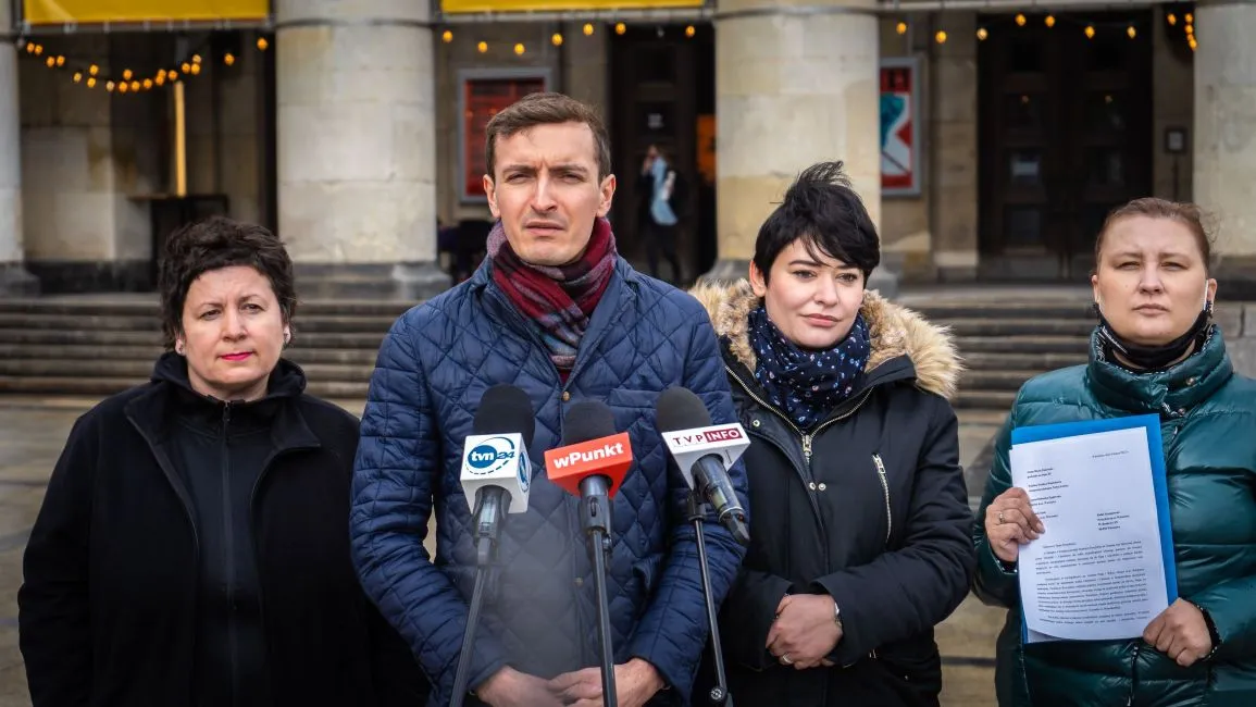 Cztery osoby stoją przed wejściem do budynku i udzielają konferencji prasowej z kilkoma mikrofonami, a jedna z nich trzyma dokument.