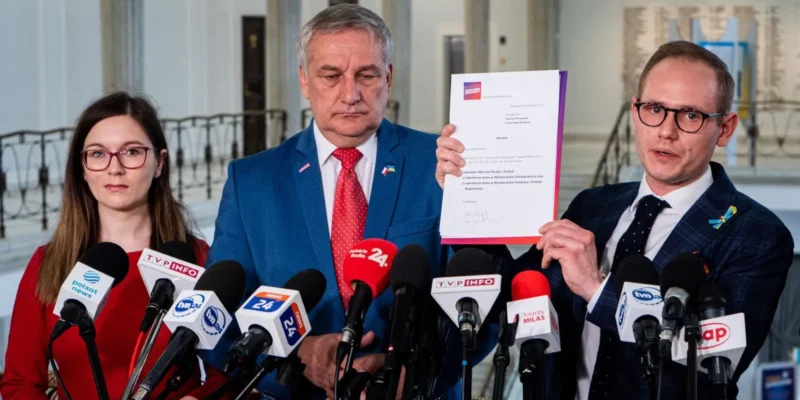 Trzy osoby na konferencji prasowej; mężczyzna w niebieskim garniturze z czerwonym krawatem trzyma dokument, obok niego drugi mężczyzna w okularach go prezentuje, a po lewej stoi kobieta w czerwonej bluzce, przed licznymi mikrofonami.