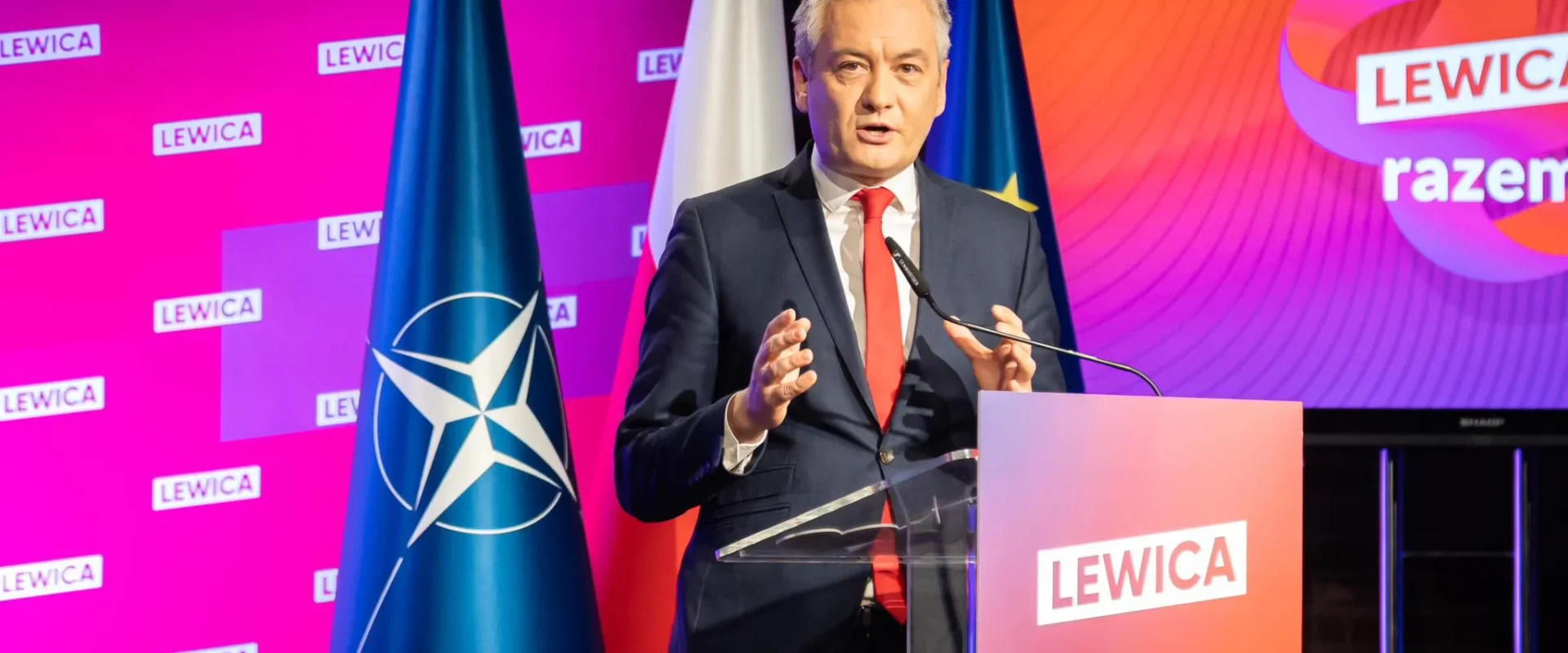 Mężczyzna w garniturze wygłasza przemówienie na scenie Lewicy, stojąc przy mównicy z logiem LEWICA; w tle widoczne flagi i kolorowy ekran z napisem LEWICA.
