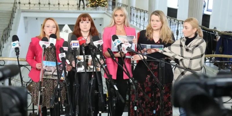 W tle choinki świątecznej, pięć kobiet w formalnych strojach prowadzi konferencję prasową, trzymając zdjęcia i stojąc przed mikrofonami.