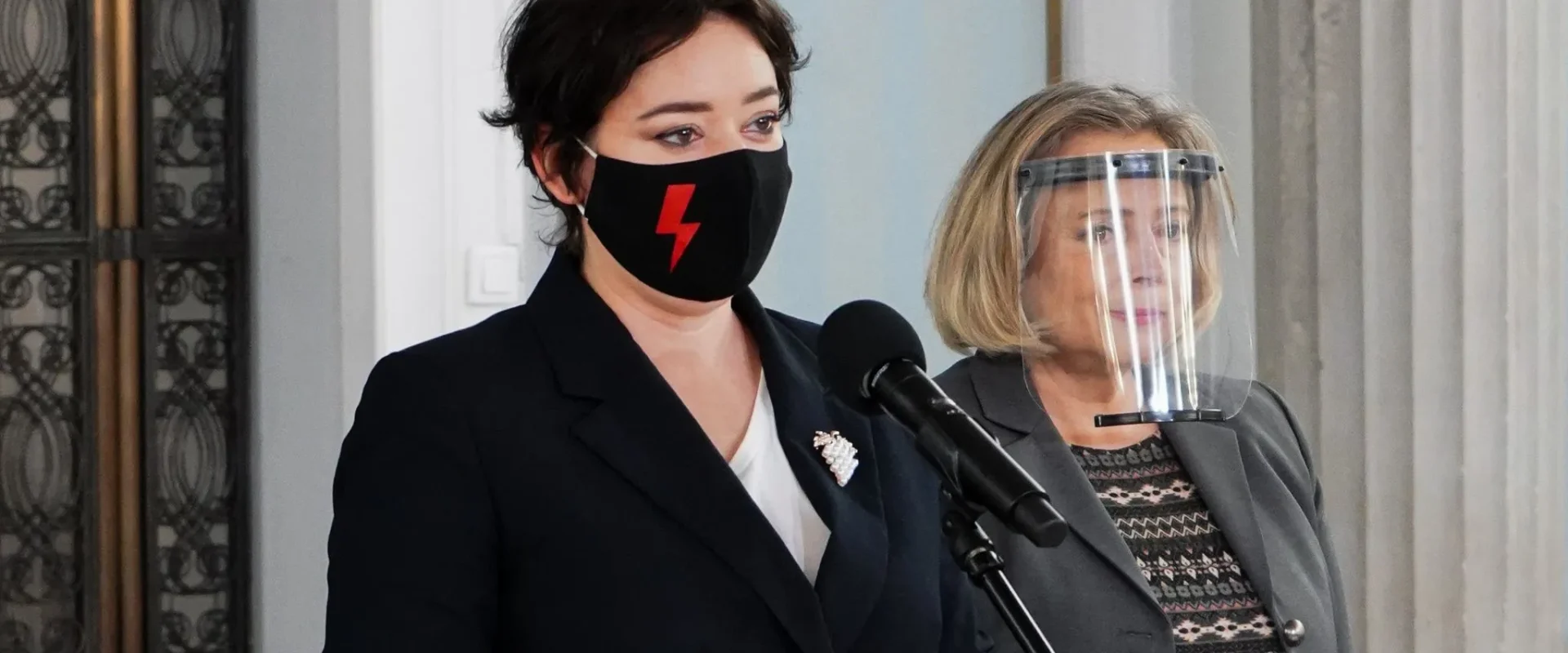 Dwie kobiety stojące przy mikrofonie podczas konferencji prasowej; po lewej w czarnej masce z czerwonym piorunem, po prawej w garniturze i z przyłbicą ochronną na twarzy.