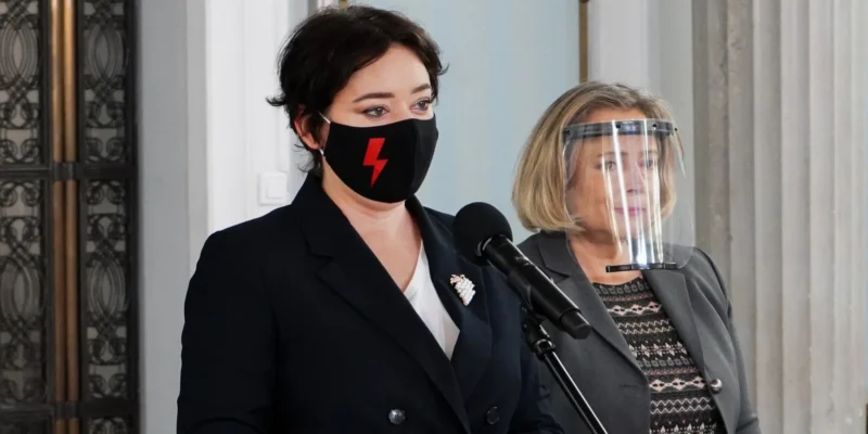 Dwie kobiety stojące przy mikrofonie podczas konferencji prasowej; po lewej w czarnej masce z czerwonym piorunem, po prawej w garniturze i z przyłbicą ochronną na twarzy.