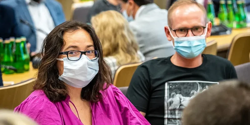 Na zdjęciu widać dwoje osób z maseczkami, siedzących w sali, w tle można dostrzec butelki wody.