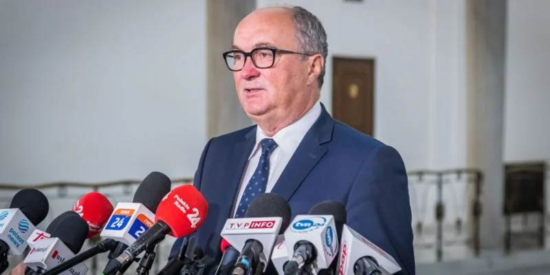 Na zdjęciu widoczny jest mężczyzna w garniturze, który udziela wywiadu, otoczony przez mikrofony telewizyjne i radiowe.