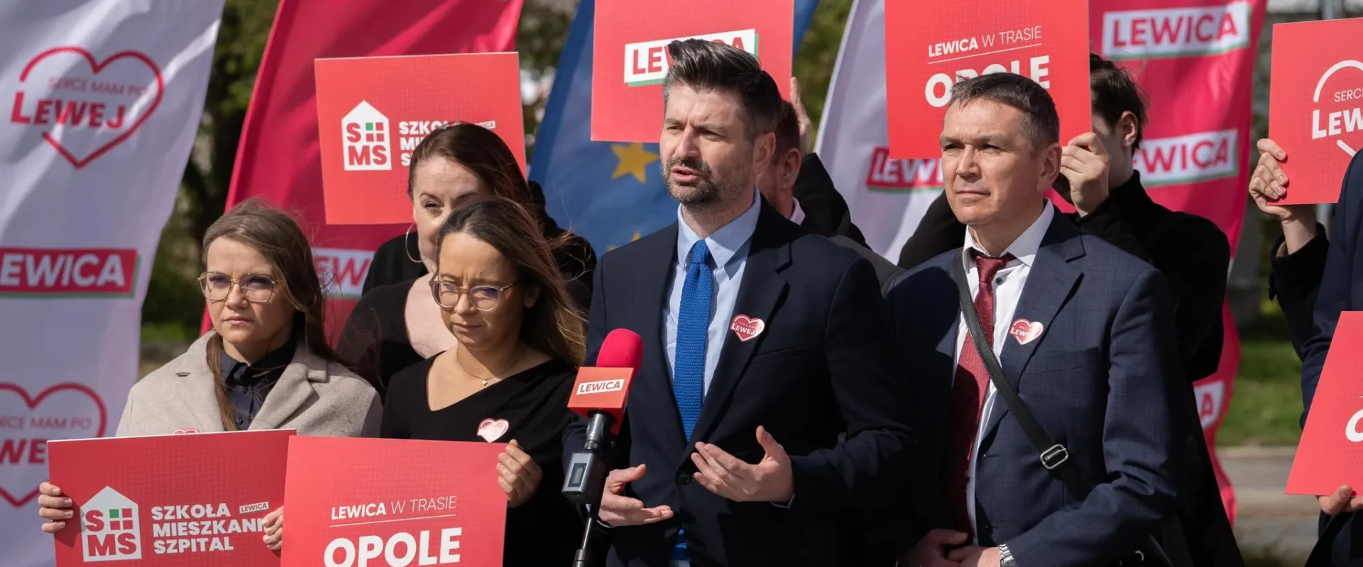 Grupa polityków Lewicy podczas konferencji prasowej na tle czerwonych banerów i plakatów z napisem LEWICA, trzyma plakaty i przemawia do mikrofonu.