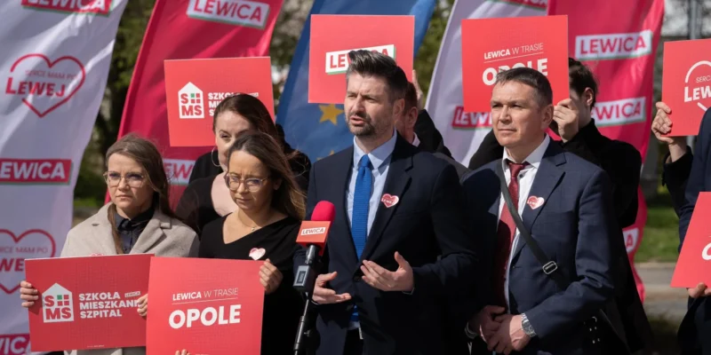 Grupa polityków Lewicy podczas konferencji prasowej na tle czerwonych banerów i plakatów z napisem LEWICA, trzyma plakaty i przemawia do mikrofonu.