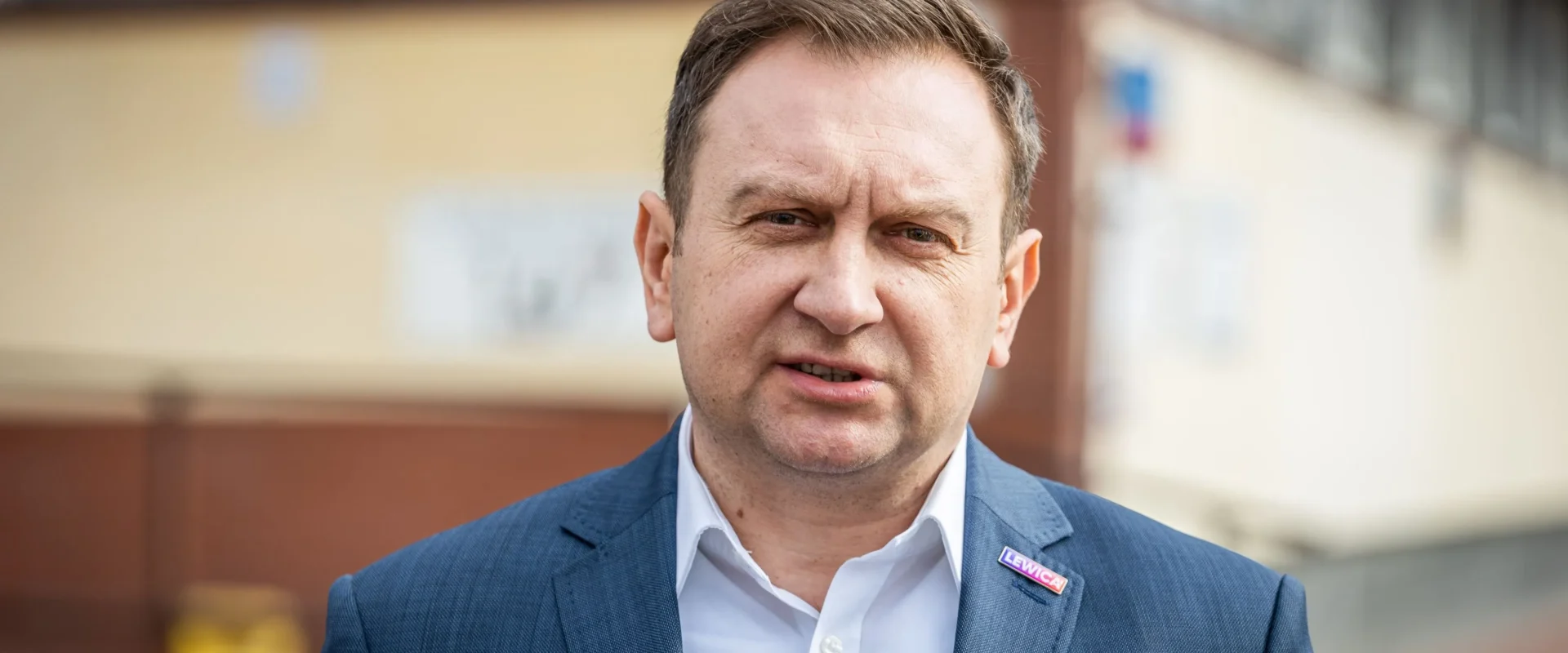 Portret mężczyzny w granatowym garniturze i białej koszuli z przypinką Lewica, stojącego na zewnątrz przy budynku.