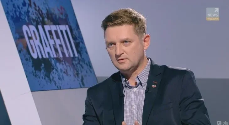 Mężczyzna w marynarce i koszuli rozmawia w studiu telewizyjnym, stojąc przed planszą z napisem „GRAFFITI”.