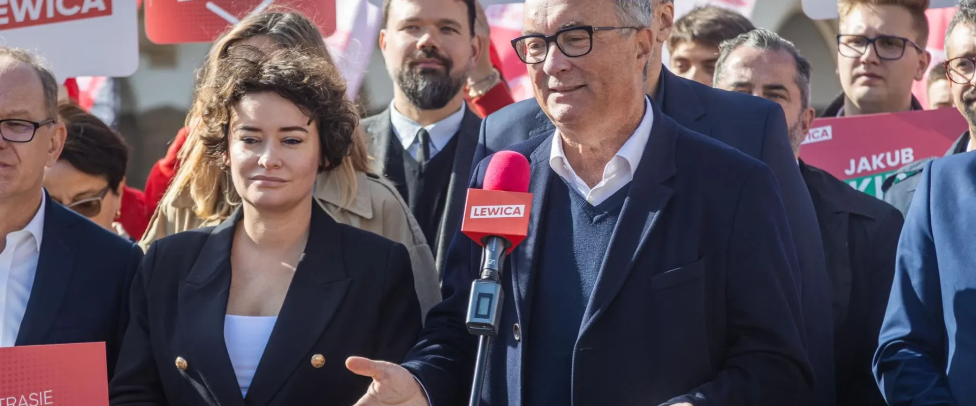 Polityk Lewicy w garniturze i okularach przemawia do mikrofonu z logiem LEWICA podczas wiecu, otoczony przez zwolenników i plakaty z hasłem LEWICA.