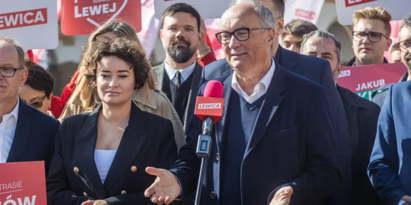 Polityk Lewicy w garniturze i okularach przemawia do mikrofonu z logiem LEWICA podczas wiecu, otoczony przez zwolenników i plakaty z hasłem LEWICA.