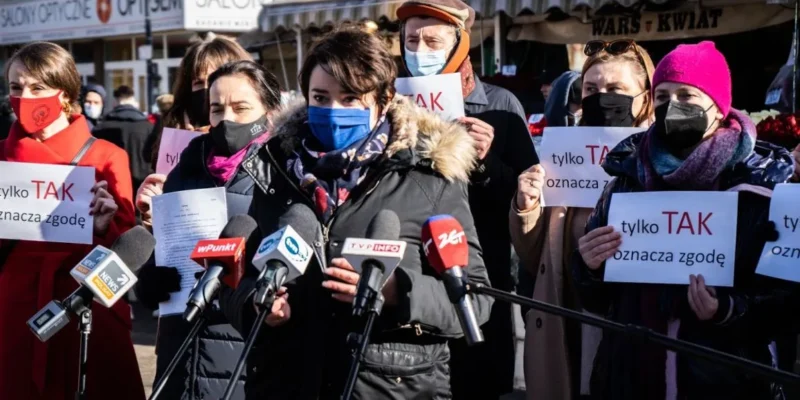 Grupa osób na konferencji prasowej trzyma plakaty z napisem „tylko TAK” i stoi wśród mikrofonów, odnosząc się do projektu zmiany definicji gwałtu.