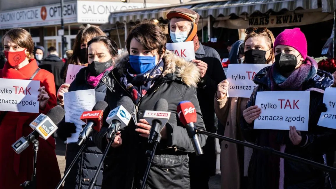 Grupa osób na konferencji prasowej trzyma plakaty z napisem „tylko TAK” i stoi wśród mikrofonów, odnosząc się do projektu zmiany definicji gwałtu.