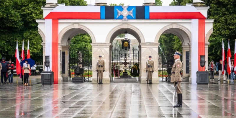 W tle architektonicznego pomnika, mundurowi stoją na baczność, a wokół rozciągają się narodowe flagi Polski podczas uroczystości w deszczowy dzień.