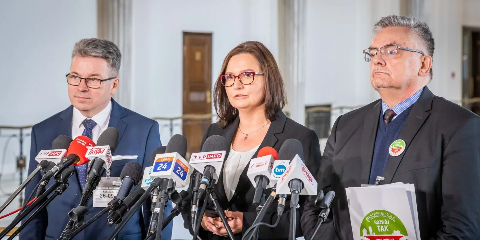 Na zdjęciu trzej mężczyźni i jedna kobieta stoją przed mikrofonami podczas konferencji prasowej, omawiając ważne kwestie społeczne.