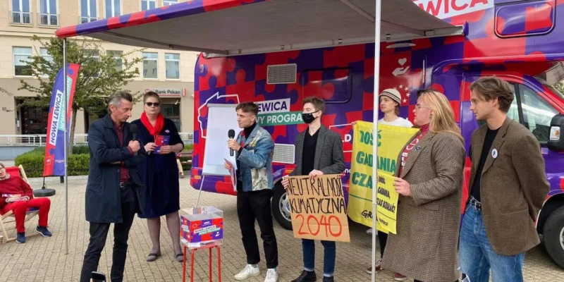 Kampania Nowej Lewicy na trasie: plenerowe wydarzenie z kamperem o jaskrawej fioletowo-czerwonej grafice, prowadzącym z mikrofonem i grupą uczestników trzymających plakaty.