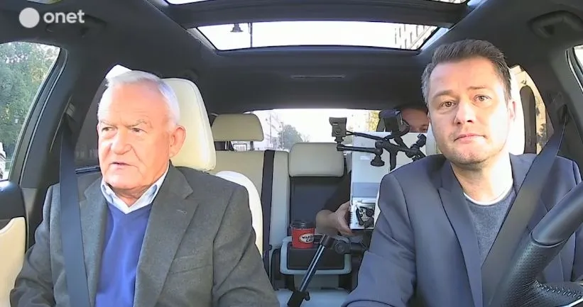 Dwaj mężczyźni siedzą w przednich fotelach samochodu podczas nagrania wywiadu, a za nimi widać kamerzystę z aparatem.