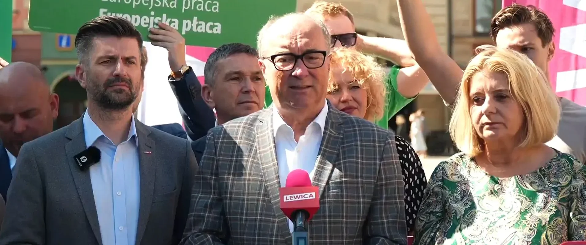 Grupa polityków i dziennikarzy podczas konferencji prasowej; centralnie stoi mężczyzna w kratkowanej marynarce, mówiący do różowego mikrofonu Lewica, w tle widoczne zielone plakaty.