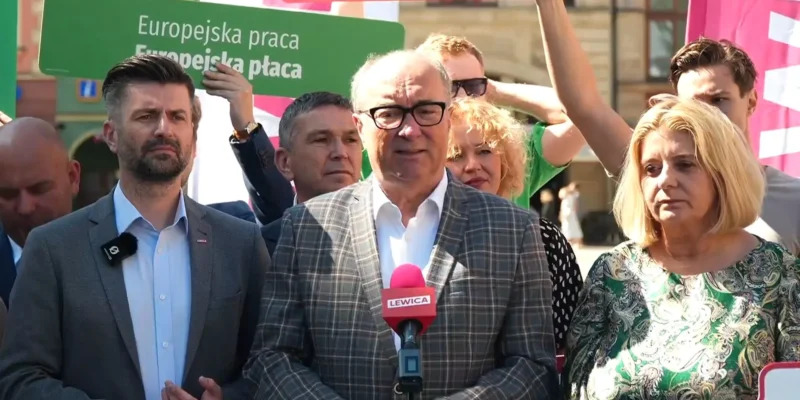 Grupa polityków i dziennikarzy podczas konferencji prasowej; centralnie stoi mężczyzna w kratkowanej marynarce, mówiący do różowego mikrofonu Lewica, w tle widoczne zielone plakaty.