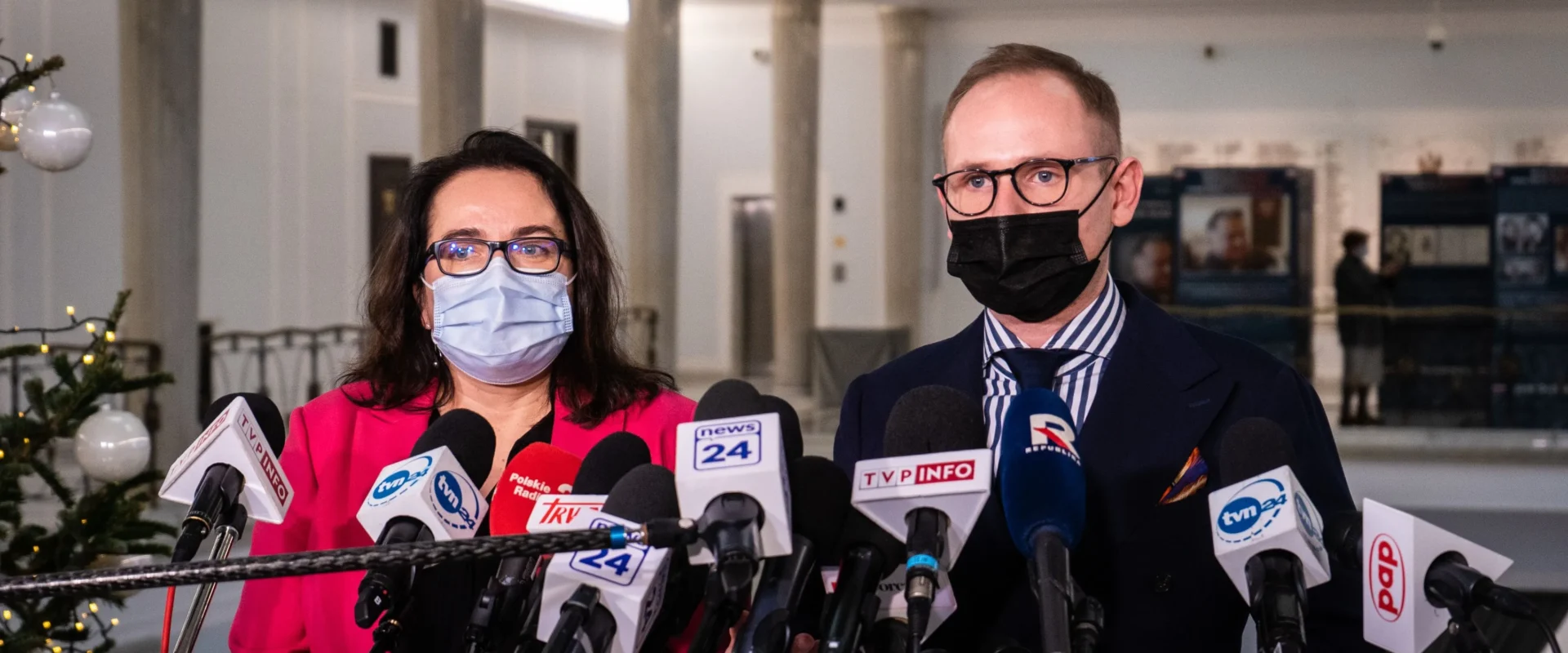 Dwóch polityków w maseczkach stoi przed licznymi mikrofonami podczas konferencji prasowej.