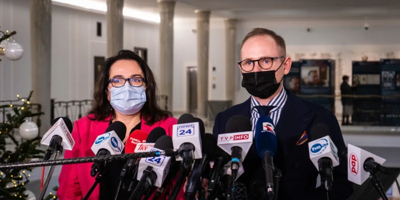 Dwóch polityków w maseczkach stoi przed licznymi mikrofonami podczas konferencji prasowej.