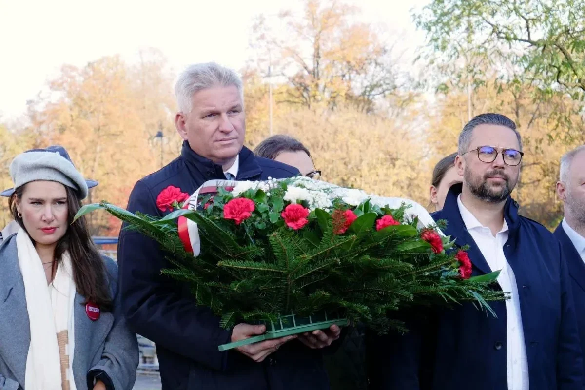 Uczestnicy ceremonii niosą duży wieniec z czerwonych i białych kwiatów podczas obchodów 105. rocznicy powołania rządu lubelskiego na tle jesiennego parku.