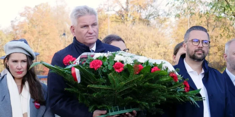 Uczestnicy ceremonii niosą duży wieniec z czerwonych i białych kwiatów podczas obchodów 105. rocznicy powołania rządu lubelskiego na tle jesiennego parku.