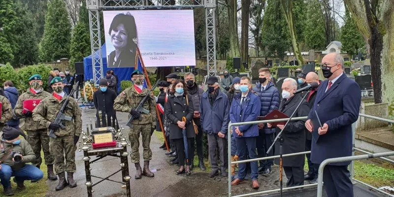 Ceremonia pożegnania na cmentarzu: żołnierze w mundurach stoją obok trumny, a zgromadzeni w maseczkach słuchają przemówienia, podczas gdy w tle na dużym ekranie wyświetlany jest portret zmarłej.
