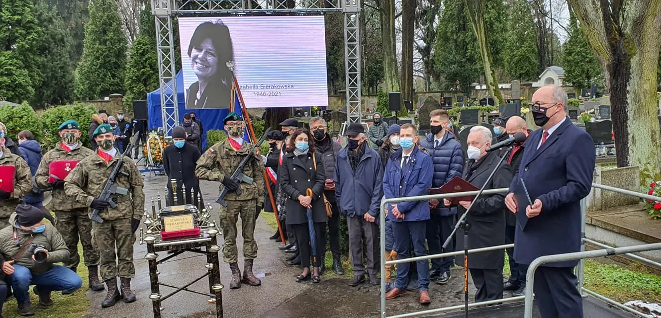 Ceremonia pożegnania na cmentarzu: żołnierze w mundurach stoją obok trumny, a zgromadzeni w maseczkach słuchają przemówienia, podczas gdy w tle na dużym ekranie wyświetlany jest portret zmarłej.