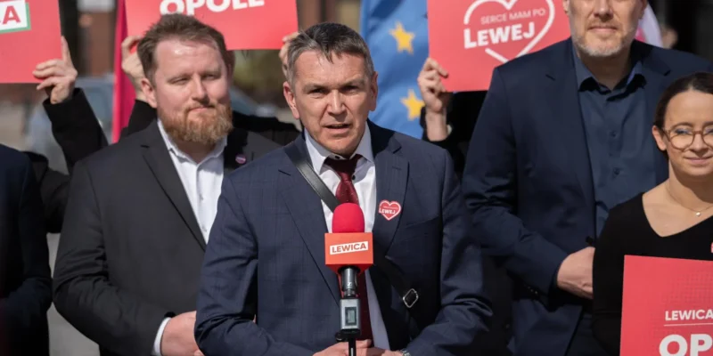 Grupa polityków Lewicy podczas konferencji prasowej, stojąca za mikrofonem z logiem Lewicy i trzymająca czerwone plakaty promujące darmowe posiłki dla uczniów.