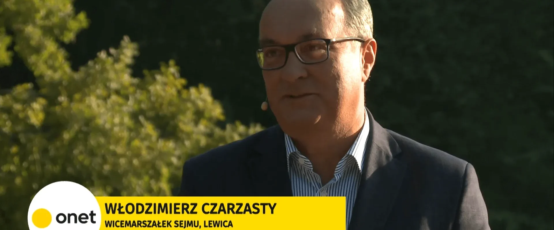 Mężczyzna w garniturze i okularach prowadzi wywiad na świeżym powietrzu na tle zieleni; na dole ekranu widnieje żółny baner z logo Onet i informacją o polityku Lewicy.