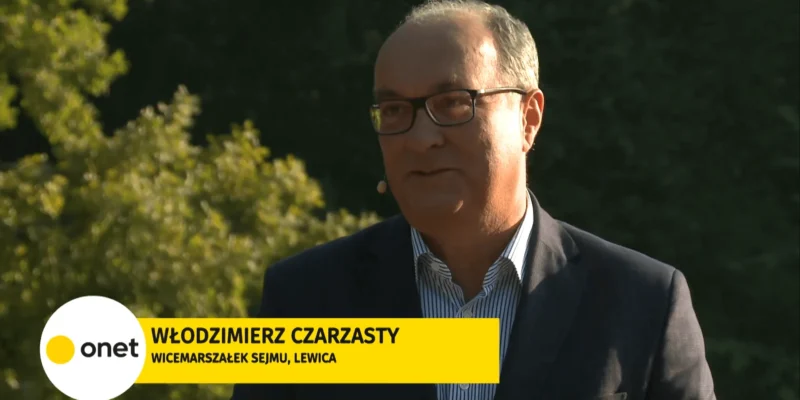 Mężczyzna w garniturze i okularach prowadzi wywiad na świeżym powietrzu na tle zieleni; na dole ekranu widnieje żółny baner z logo Onet i informacją o polityku Lewicy.