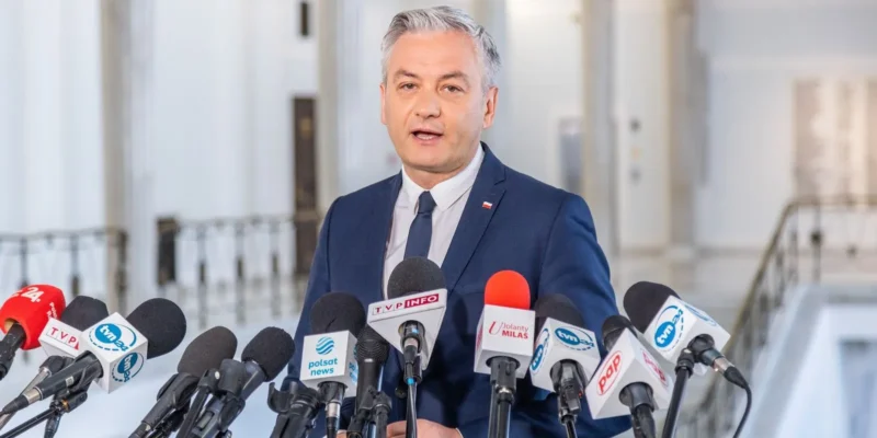 Mężczyzna w granatowym garniturze wygłasza oświadczenie podczas konferencji prasowej, otoczony licznymi mikrofonami dziennikarskimi.