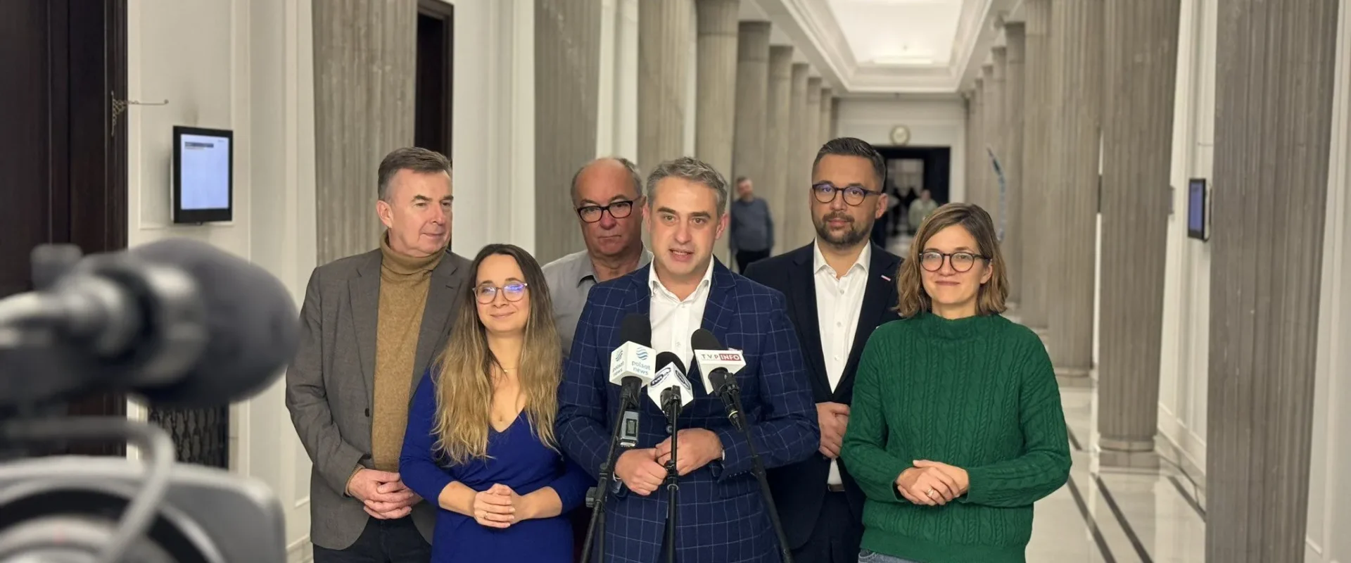 Grupa polityków Koalicyjnego Klubu Parlamentarnego Lewicy stoi w korytarzu przed mikrofonami podczas konferencji prasowej.