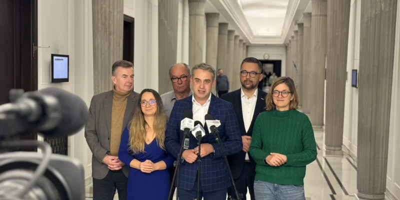 Grupa polityków Koalicyjnego Klubu Parlamentarnego Lewicy stoi w korytarzu przed mikrofonami podczas konferencji prasowej.