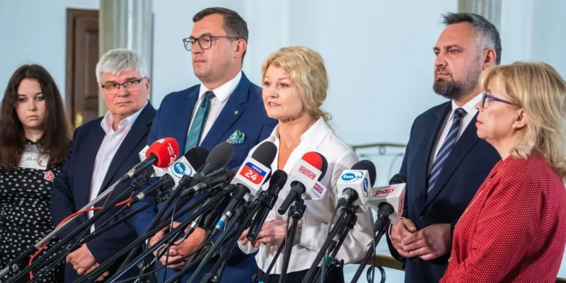 Na zdjęciu widać grupę siedmiu osób podczas konferencji prasowej, stojących przed mikrofonami z różnymi logotypami mediów.