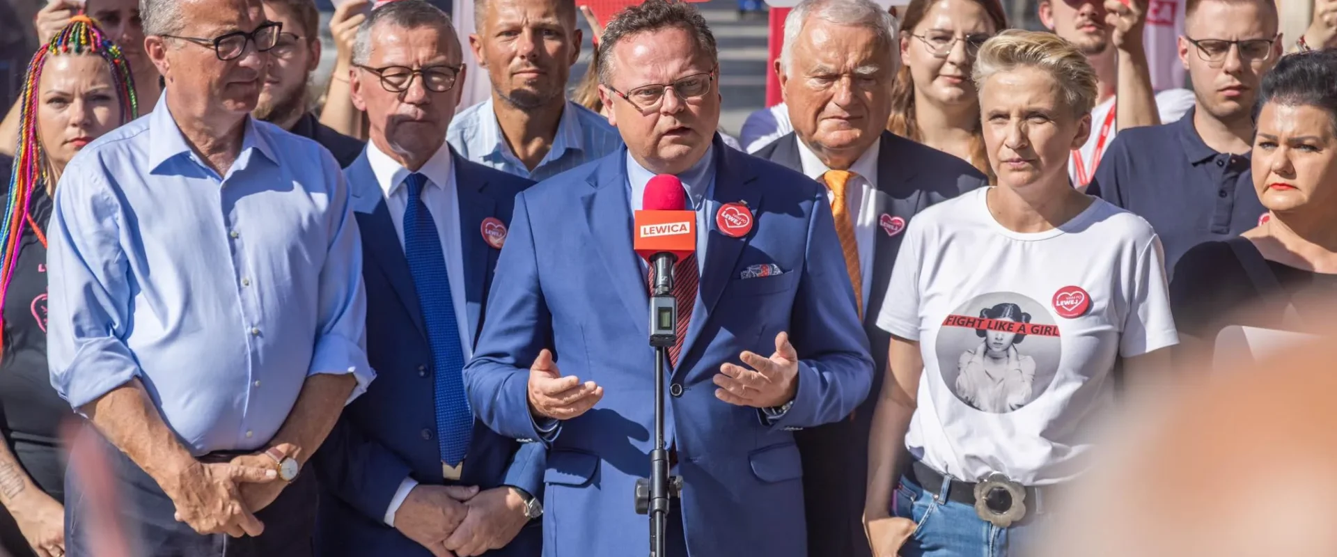 Grupa zwolenników Lewicy podczas konferencji prasowej na tle plakatów LEWICA, centralnie stojący mężczyzna w granatowym garniturze przemawia do mikrofonu, nawiązując do kontroli na granicy polsko‑niemieckiej.