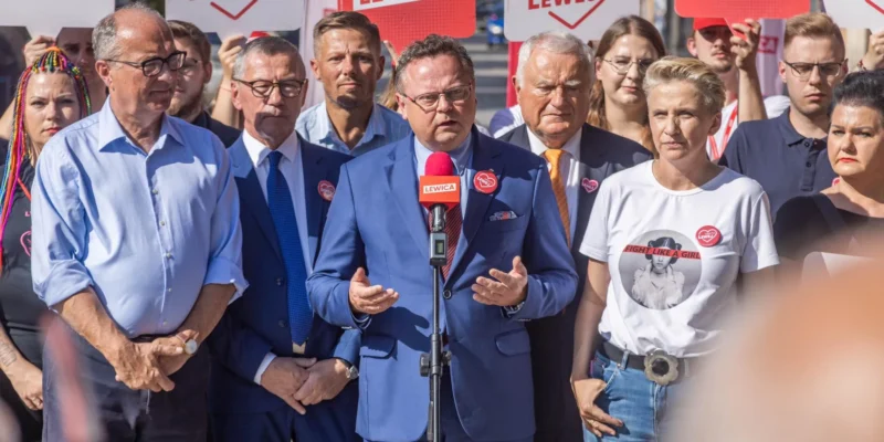 Grupa zwolenników Lewicy podczas konferencji prasowej na tle plakatów LEWICA, centralnie stojący mężczyzna w granatowym garniturze przemawia do mikrofonu, nawiązując do kontroli na granicy polsko‑niemieckiej.