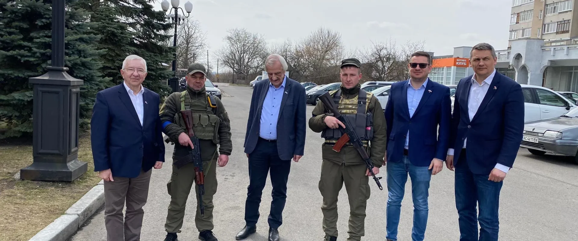 Sześć mężczyzn stoi na ulicy w Łucku; dwie osoby w mundurach wojskowych z bronią, pozostali w garniturach, w tle drzewa i budynki.