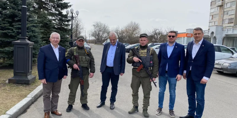 Sześć mężczyzn stoi na ulicy w Łucku; dwie osoby w mundurach wojskowych z bronią, pozostali w garniturach, w tle drzewa i budynki.