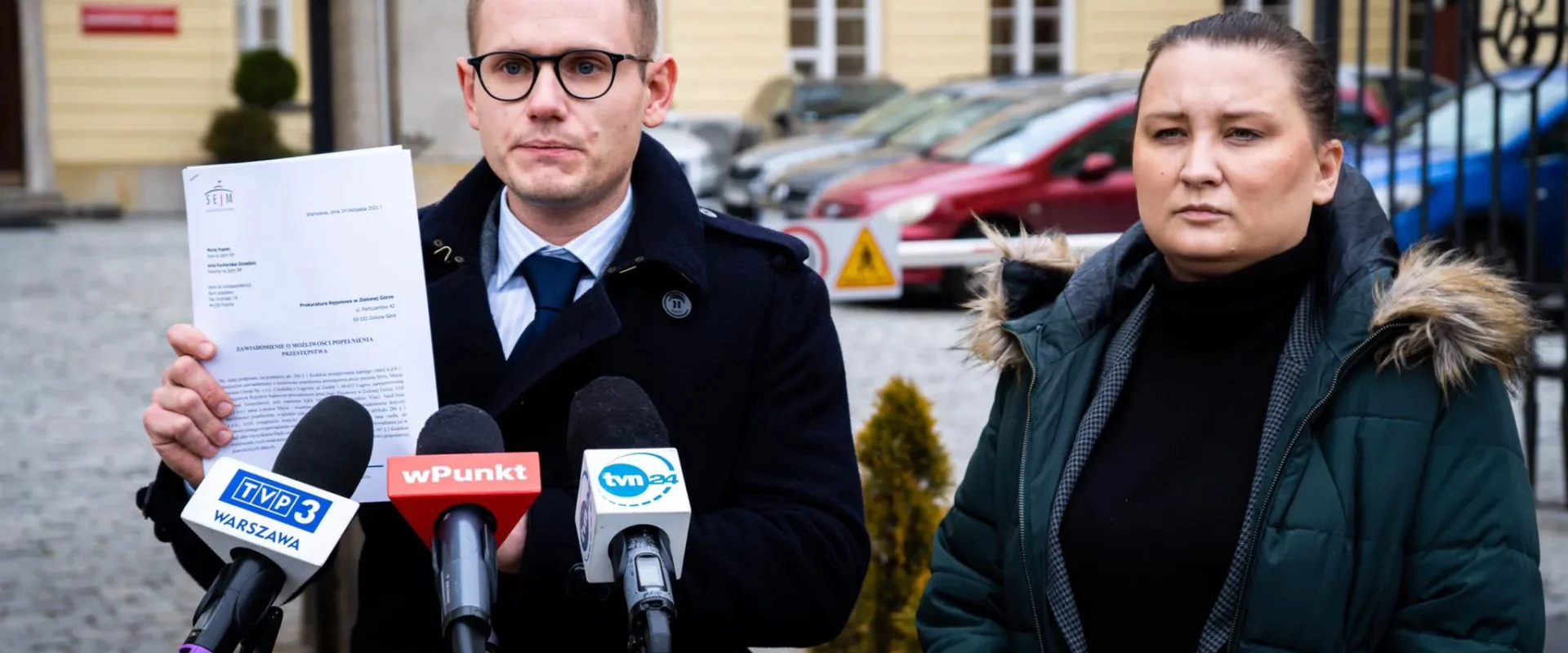 Na zdjęciu widoczni są mężczyzna w okularach trzymający dokument oraz kobieta stojąca obok niego, podczas konferencji prasowej na tle budynku.