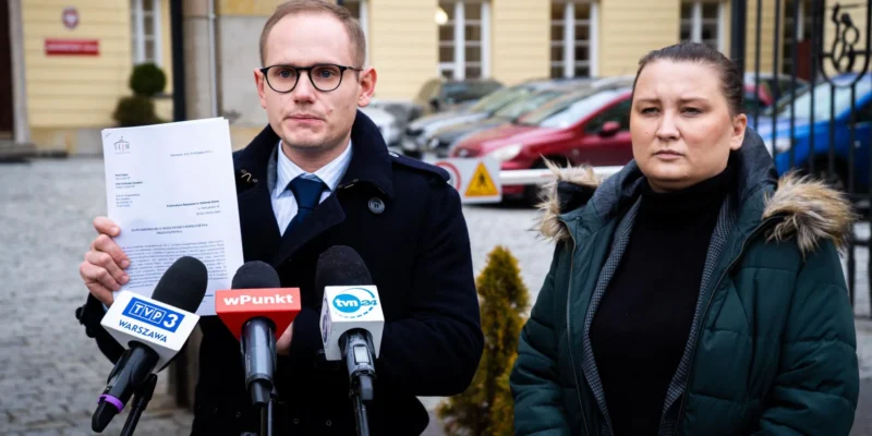 Na zdjęciu widoczni są mężczyzna w okularach trzymający dokument oraz kobieta stojąca obok niego, podczas konferencji prasowej na tle budynku.